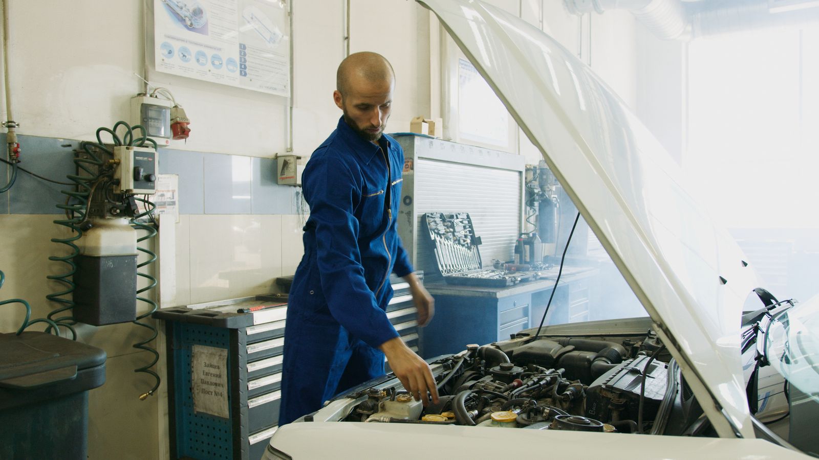 Mechanic in a blue uniform inspecting the engine of a car with the hood open in a repair shop