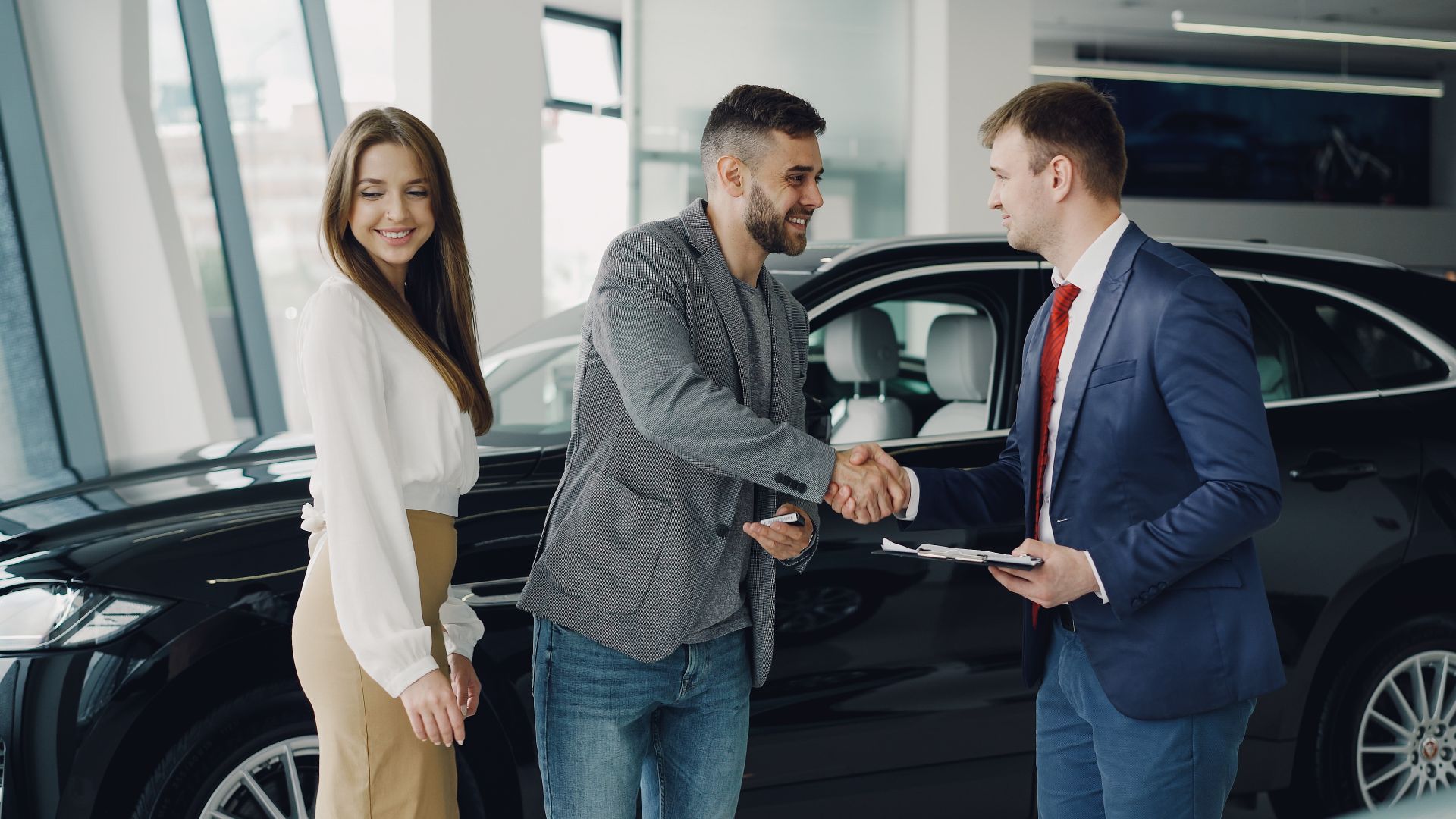 Car buyer shaking hands with a salesperson holding paperwork beside a black SUV in a dealership showroom