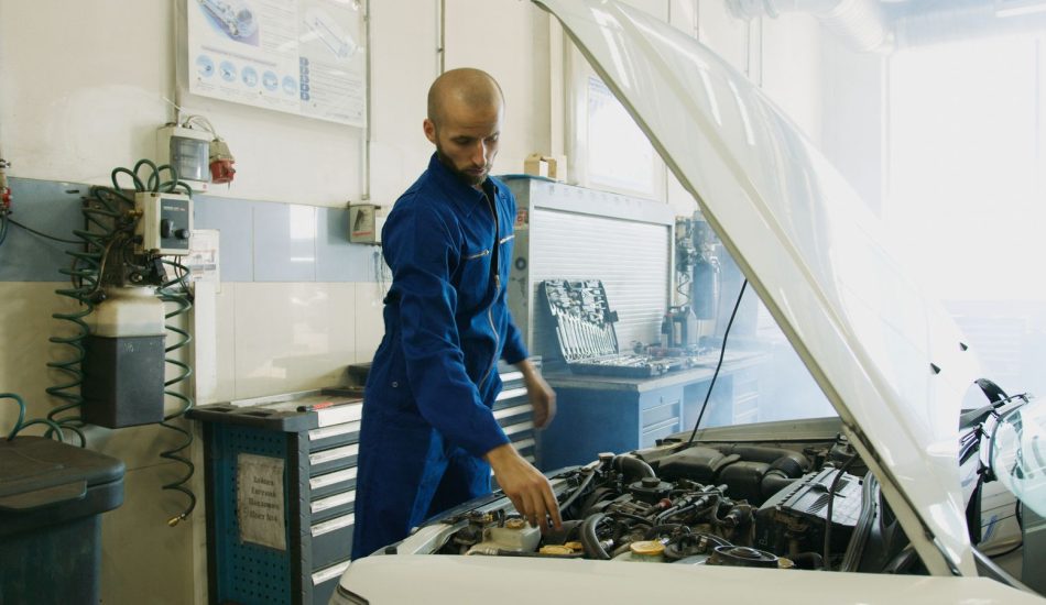 Mechanic in a blue uniform inspecting the engine of a car with the hood open in a repair shop
