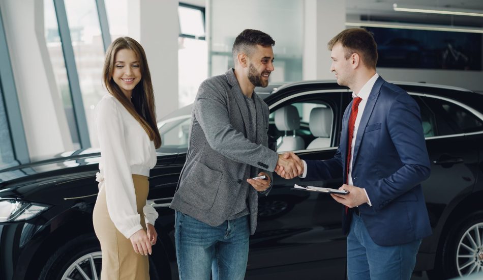 Car buyer shaking hands with a salesperson holding paperwork beside a black SUV in a dealership showroom